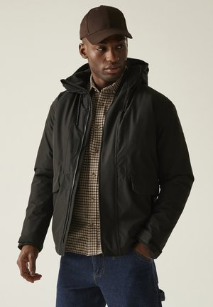 Mężczyzna w czarnej zimowej kurtce z kapturem, brązowej koszuli w kratę, niebieskich dżinsach i ciemnobrązowej czapce, stojący z jedną ręką w kieszeni.