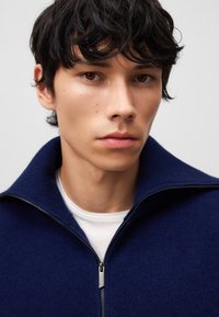 Maglione blu navy con colletto alto e chiusura con zip, realizzato in tessuto morbido. Indossato sopra una camicia bianca a girocollo, con dettagli di design minimi.