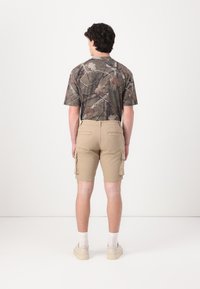 T-shirt camouflage à manches courtes, associé à un short cargo kaki avec poches latérales ; le mannequin porte des baskets claires et des chaussettes blanches.