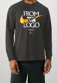 Black long-sleeve shirt made of cotton featuring bold white and orange Nike logo text, with “FROM THE LOGO” prominently displayed.
