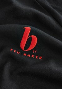 Čierna tkanina s hladkým povrchom, na ktorej je výrazné červené vyšívané logo "b" nad menším textom "TED BAKER".
