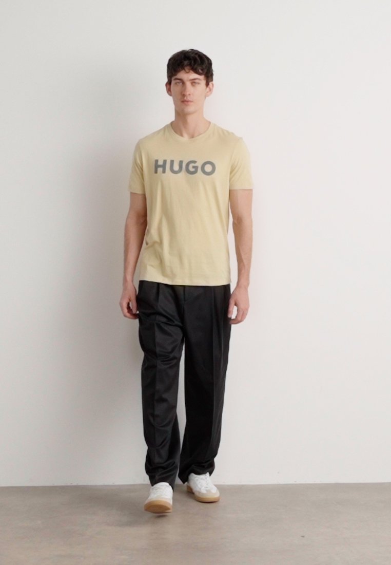 Светло-жълта тениска с логото "HUGO" в сиво, съчетана с черни, широки панталони и бели маратонки. Минималистичен дизайн, удобна кройка.