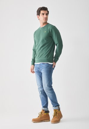 Groene sweatshirt met een ronde hals, gecombineerd met lichtblauwe denim jeans en bruine werkschoenen. Opvallend geel logo op de sweatshirt.