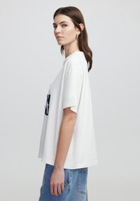 Witte oversized katoenen t-shirt met korte mouwen met een donkerblauwe graphic op de voorkant. Gemaakt met lichte blauwe denim jeans.
