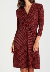 Robe à motifs rouge et noir avec un décolleté en V, des manches trois-quarts et un détail de taille nouée, fabriquée à partir d'un tissu doux et texturé.