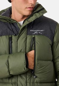 Green puffer jacket featuring a black panel with a grid pattern, zippered pockets, and logo, designed for warmth and functionality.