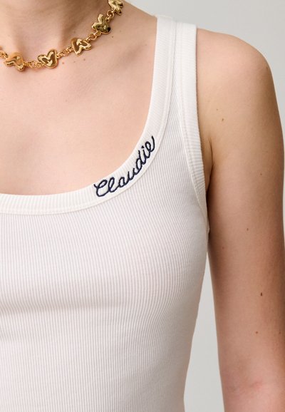 Hombro y parte superior del pecho con una camiseta de tirantes blanca con canalé con "Claudie" bordado cerca del escote y un collar de cadena dorada con mariposa.