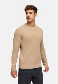 Maglione a coste beige con scollatura rotonda e maniche lunghe, abbinato a jeans scuri. La texture sembra morbida e calda.