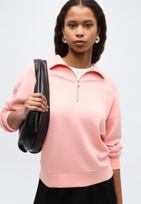 Νεαρή γυναίκα που φοράει ροζ φούτερ με φερμουάρ στο ένα τέταρτο και χρυσούς κρίκους στα αυτιά, κρατώντας μια μαύρη δερμάτινη τσάντα ώμου μπροστά σε απλό φόντο.