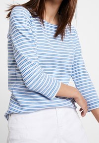 Haut à manches longues rayé présentant des lignes horizontales bleues et blanches, en tissu doux, avec un col rond et des fentes sur les côtés pour un confort de mouvement.