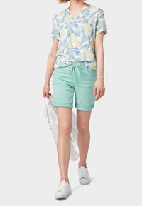 Camisa de manga corta con un patrón de hojas de palma amarillas, azules y verdes, combinada con pantalones cortos verde menta y zapatillas blancas.