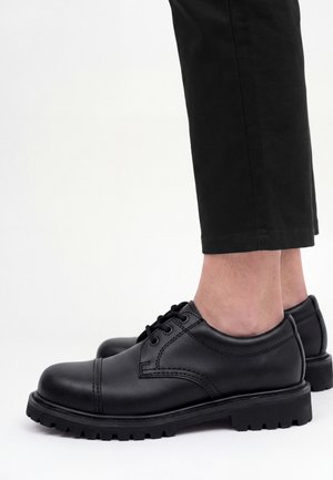 Personne portant des chaussures en cuir noires à lacets avec des semelles épaisses et un pantalon noir court sur fond blanc.