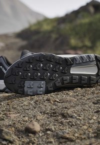 Close-up of a black and gray hiking shoe sole with deep treads resting on rocky, uneven ground outdoors.