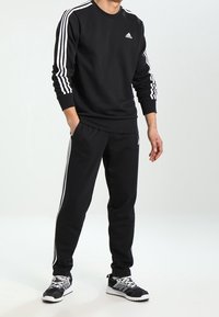 Czarny zestaw z bluzy i spodni z miękkiego materiału, z białymi akcentami w postaci trzech pasków na rękawach i bokach, połączony z czarnymi sneakersami.