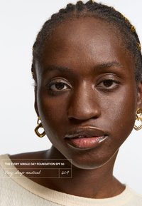 Every Single Day Foundation SPF 50 sävyssä Very Deep Neutral 609. Lähikuva mallin kasvoista, joilla on punottu hius ja kultaketjut korvissa, esittelee sileää ihotekstuuria.