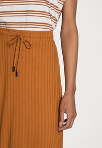 Pantalones acanalados de color óxido con cintura elástica y cordón ajustable. Textura suave con pequeños extremos metálicos negros en el cordón.