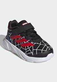 Baskets de sport noires avec un motif de toile d'araignée blanche, des accents rouges et une sangle Velcro. Présente une semelle en caoutchouc texturée et un dessus en maille respirante.