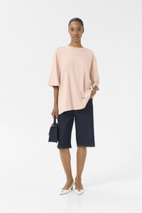 Lys rosa oversized t-skjorte, denim bermudashorts, hvite hæler med liten blokkhel, og en liten marineblå håndveske.