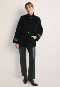 Czarna polarowa kurtka z wysokim kołnierzem, z przodu zapinana na guziki i z kieszeniami po bokach; czarne skórzane akcenty na rękawach. Połączona z czarnymi skórzanymi spodniami i obcasami.