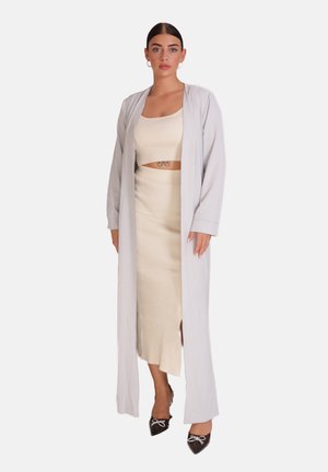 Un long cardigan gris clair porté sur un top court à côtes beige et une jupe longue. La tenue présente une fente et des escarpins noirs à talon pointu avec un accent de nœud.