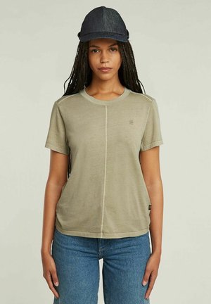 Donna con capelli lunghi intrecciati che indossa una maglietta beige a maniche corte, jeans blu e un berretto da baseball grigio scuro, in piedi davanti a uno sfondo neutro.