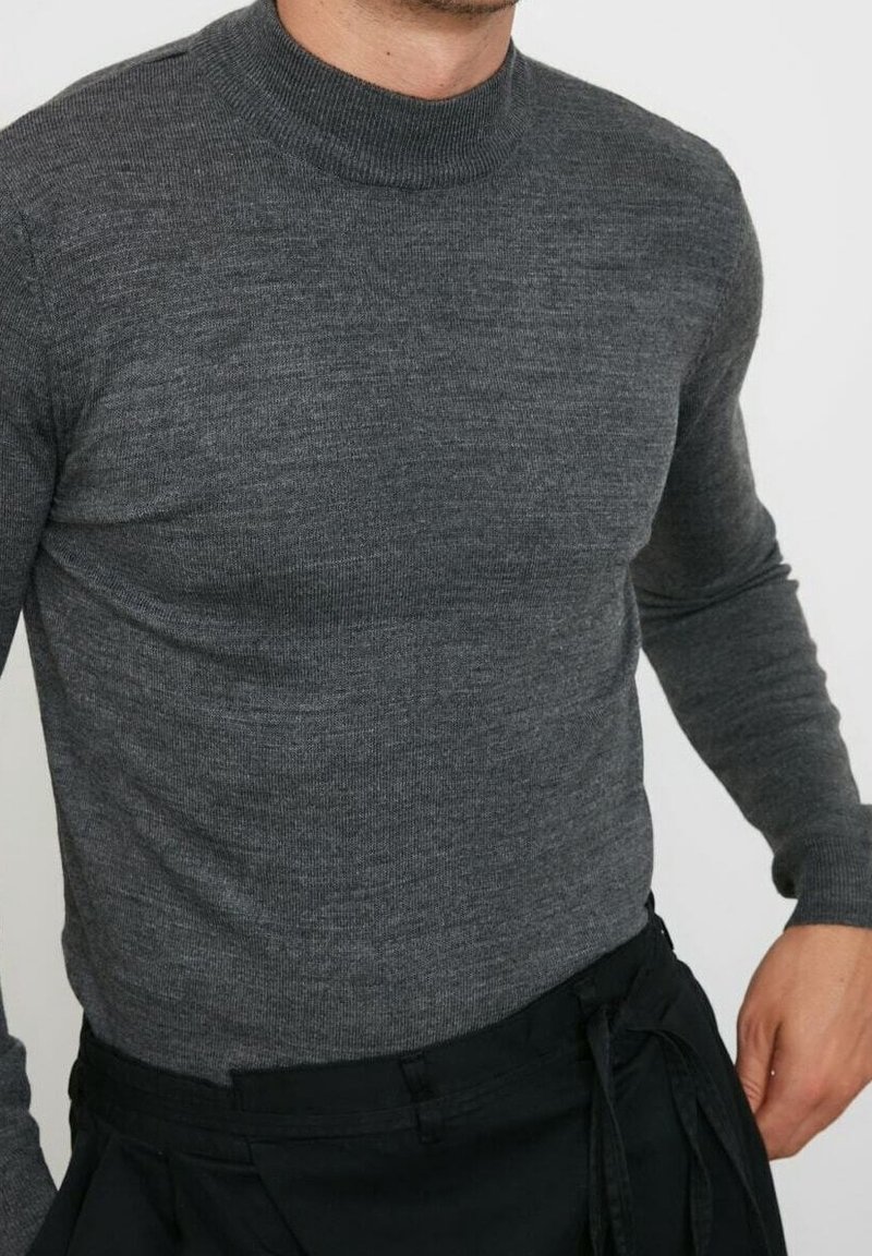 Homme portant un pull en maille gris foncé cintré avec un col montant et un pantalon noir, debout devant un fond uni.