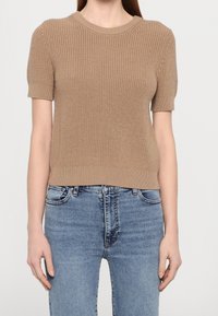 Kortärmad stickad tröja i beige med ribbad textur, rund halsringning och figurnära silhuett, kombinerad med ljusblå jeans.