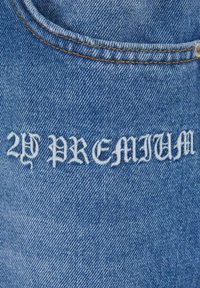 Denim tkanina, svijetloplava, s natpisom "2Y PREMIUM" izvezenim u bijelom gotičkom fontu, s kontrastnim žutim šavovima duž rubova.