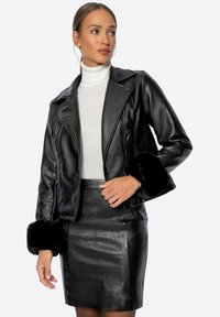 Giacca in pelle nera con polsini in pelliccia sintetica, chiusura con zip frontale e colletto a incavo, abbinata a una gonna a matita nera per un look coordinato.