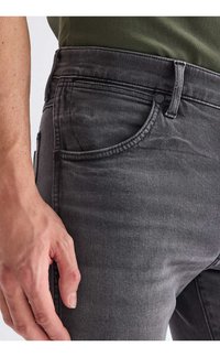 Prikaz roke osebe, ki počiva ob žepu izbledelih črnih kavbojk in zeleno srajco, ki je zataknjena noter.