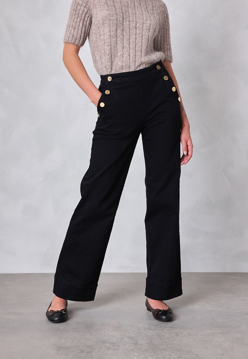 Czarne spodnie o szerokich nogawkach z złotymi guzikami przy kieszeniach, dopasowanym paskiem w talii i gładką fakturą materiału. Połączone z prążkowanym, jasnym topem.