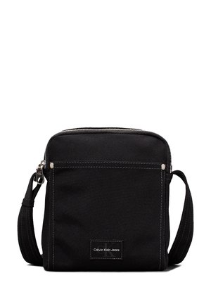 Kleine schwarze Umhängetasche mit Fronttasche, silbernem Reißverschluss, verstellbarem Riemen und Calvin Klein Jeans Logo-Patch auf der Vorderseite.