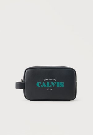 Trousse de toilette rectangulaire noire avec logo « Calvin Klein » bleu sarcelle et poignée latérale sur un fond clair uni.