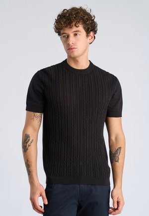 Ung mand med krøllet hår iført sort kortærmet striktrøje, der viser tatoveringer af en ørn og en sommerfugl på underarmene, som kigger til siden.