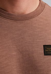 Hnědá bavlněná mikina s žebrovaným kulatým výstřihem a malou černou a žlutou etikettou vpředu, vyrobená z texturované tkaniny.