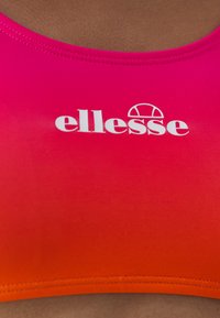 Športový top s gradientom od ružovej po oranžovú, vyrobený z hladkej tkaniny, s bielym logom "ellesse" umiestneným v strede oblasti hrudníka.