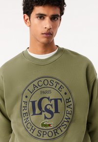 Felpa verde oliva in cotone, con un grande logo blu navy con la scritta "LACOSTE" e un accento di coccodrillo ricamato sul davanti.