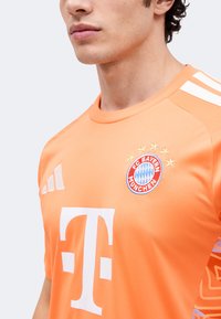 Άνδρας που φοράει πορτοκαλί φανέλα της FC Bayern München με λευκές ρίγες Adidas και λογότυπο Telekom, δείχνοντας μόνο το στήθος και την περιοχή του λαιμού.