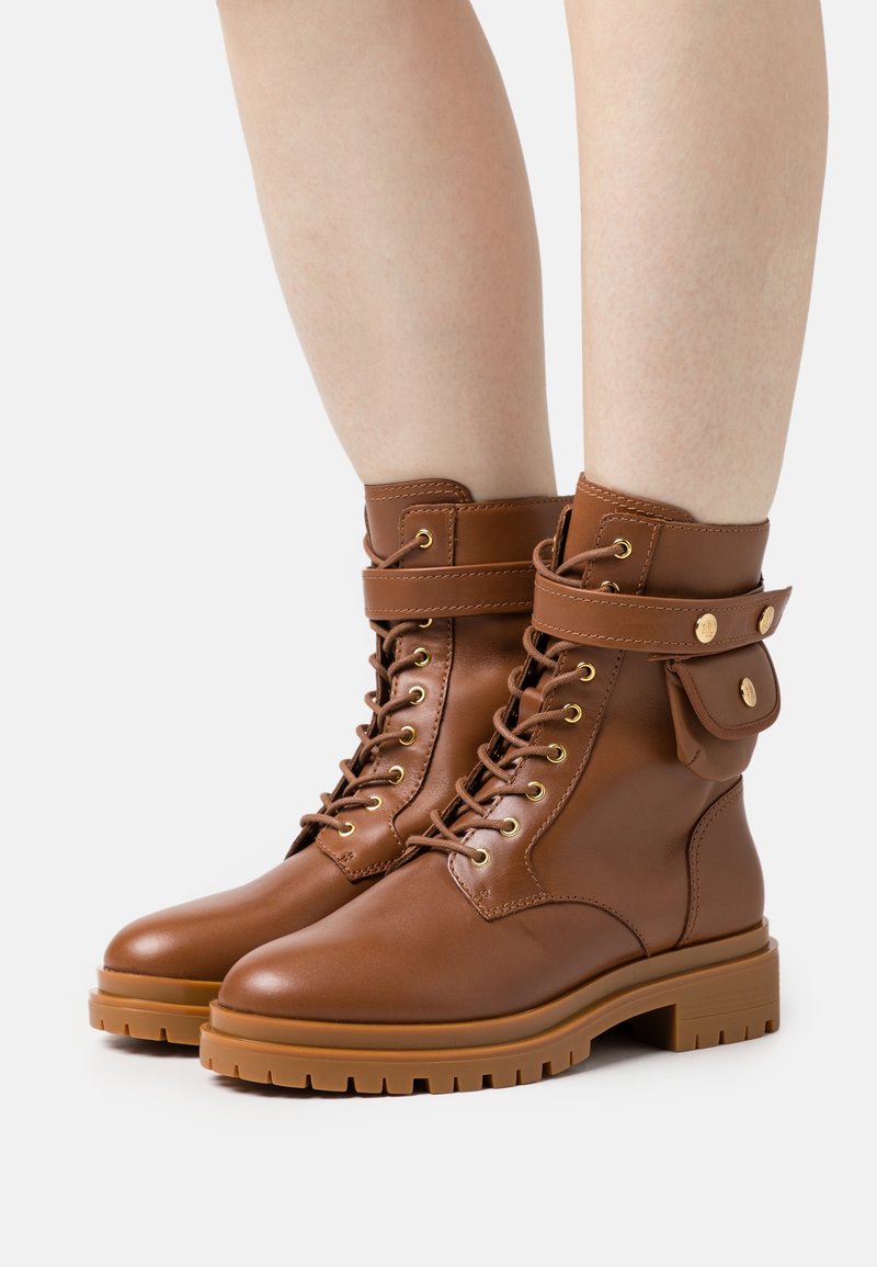 Stivali alla caviglia in cuoio marrone con lacci frontali, occhielli dorati e una fascia decorativa. Presentano una piccola tasca laterale e una suola in gomma spessa.