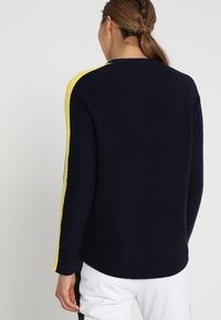 Pull en laine bleu marine avec des bandes latérales jaunes, texture côtelée, manches longues et coupe décontractée. Vue arrière mettant en valeur le contraste de couleurs.