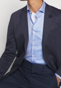 Blazer blu navy sopra una camicia azzurra chiara a fantasia con chiusura a bottoni. La camicia presenta una sottile texture a diamante. I pantaloni sono di un blu navy scuro.