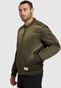 Olivgrüne Bomberjacke aus glänzendem Nylon, mit einem Frontreißverschluss, gerippten Bündchen, Seitentaschen und einem Aufnäher am Saum.