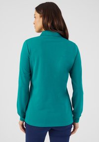 Blusa de manga larga en turquesa con cuello y silueta ajustada, fabricada con una tela suave y con puños ligeramente fruncidos. Textura suave.