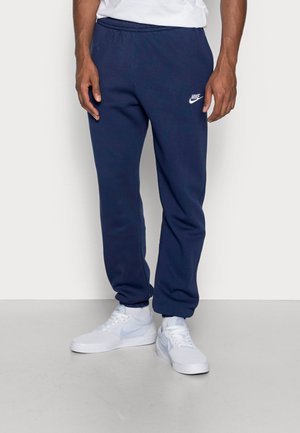 Mann, der dunkelblaue Nike-Jogginghose und weiße Nike-Sneaker trägt, steht vor einem schlichten hellen Hintergrund.