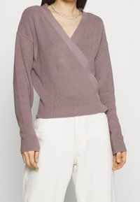 Suéter de punto ligero color rosa empolvado con escote en V y diseño cruzado. Las mangas largas tienen puños acanalados. Combinado con pantalones color crema.