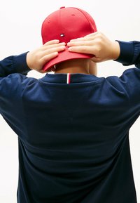 Rote Baseballmütze mit flachem Schirm, rückwärts getragen über einem marineblauen Sweatshirt, das am Kragen einen kleinen rot-weiß-blauen Streifen hat.