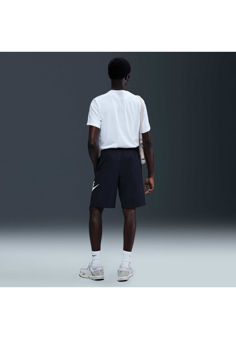 Hombre de pie de espaldas, vistiendo camiseta blanca, pantalones cortos negros con logo, calcetines blancos, zapatillas deportivas y llevando un bolso de hombro de color claro.