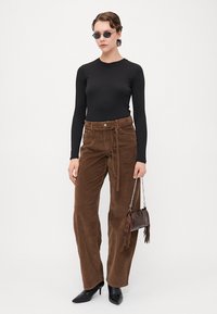 Bluză neagră cu mâneci lungi, pantaloni largi din catifea călcată maro cu un brâu legat, și tocuri negre ascuțite. Accesoriile includ o geantă mică maro.