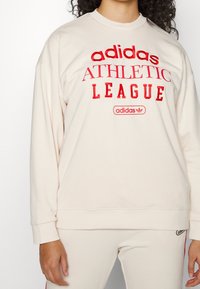 Crèmekleurige sweatshirt met gedurfde rode tekst "adidas ATHLETIC LEAGUE", met een klein logo, lange mouwen en een ontspannen pasvorm.