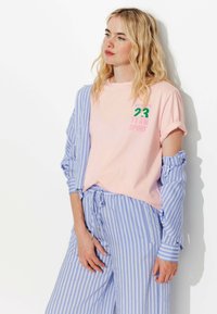 Chemise courte rose clair à manches courtes avec du texte vert et rose. Associée à un pantalon ample bleu et blanc rayé, avec une taille élastiquée.